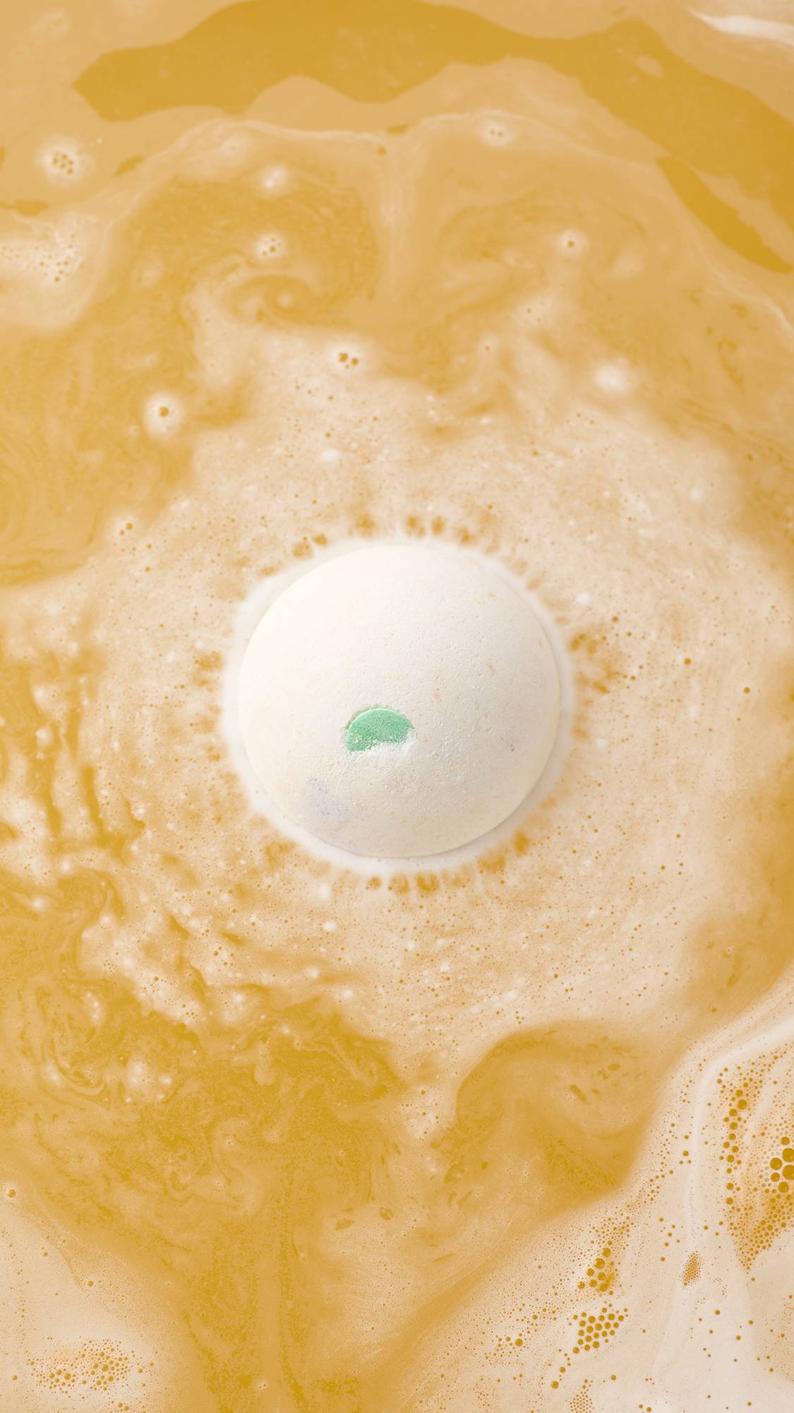 An aerial view of the "Dragon's Egg" bath bomb with a small green accent dissolving in vibrant orange water. It is surrounded by a ring of frothy white bubbles and swirling, marbled patterns across the surface.