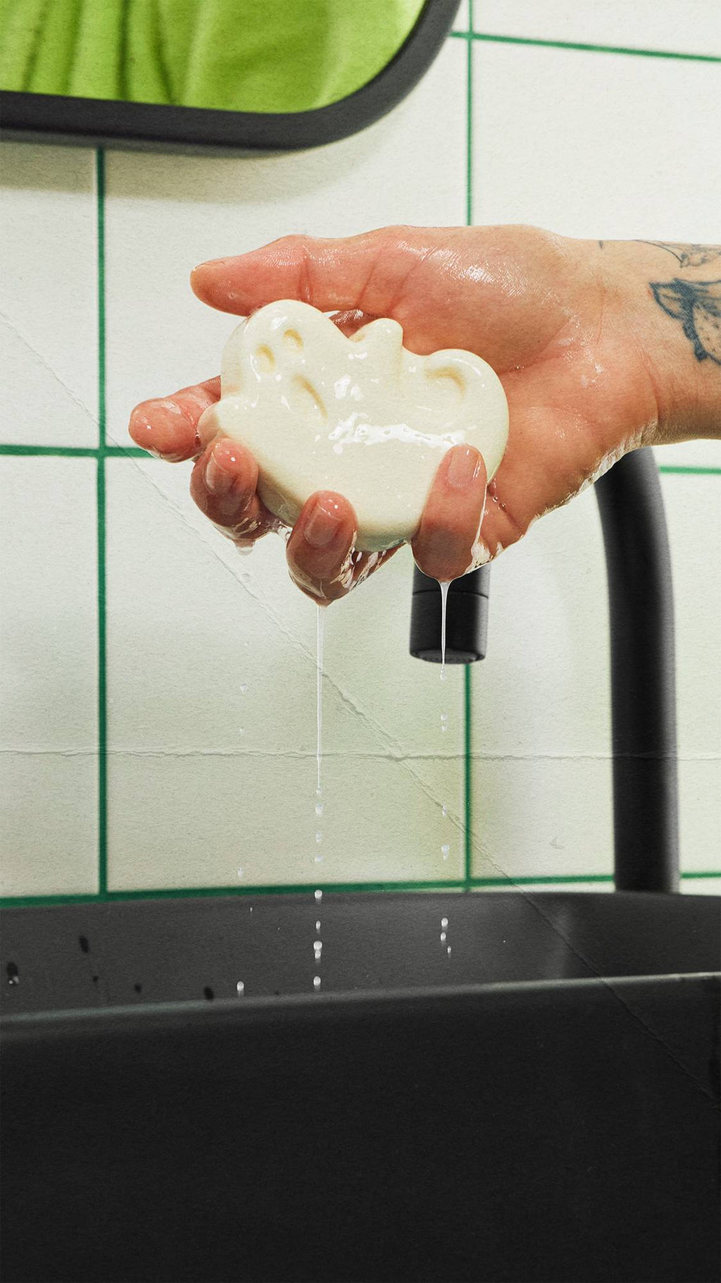 A wet, tattooed hand holds a ghost-shaped off-white bar of soap over a black sink as water drips down. A black tap and white wall tiles with green grout lines are visible in the background.
