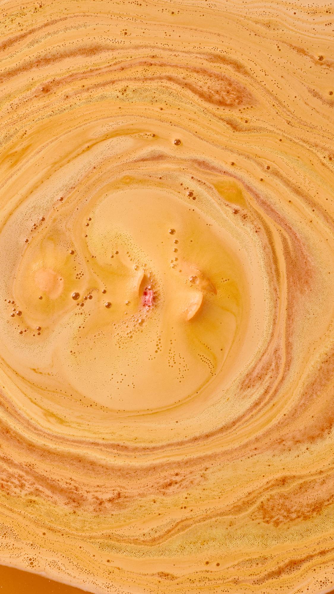 Top-down view of a dissolved Peachy Bath Bomb creating mesmerizing swirls of vibrant orange and golden foam with tiny bubble trails.