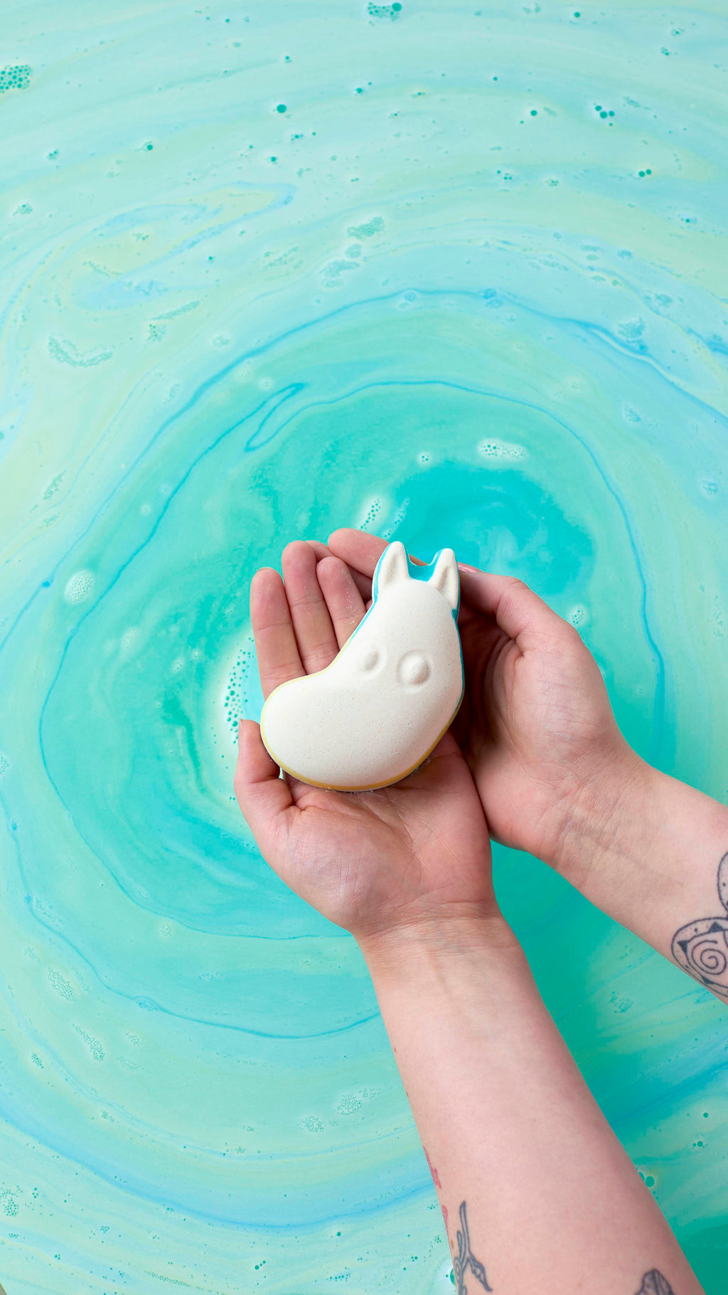 An overhead shot of two hands holding a white Moomintroll bath bomb over swirling turquoise and light green bathwater. The water creates a circular, marbled pattern around the hands, which have tattoos visible on the forearms.