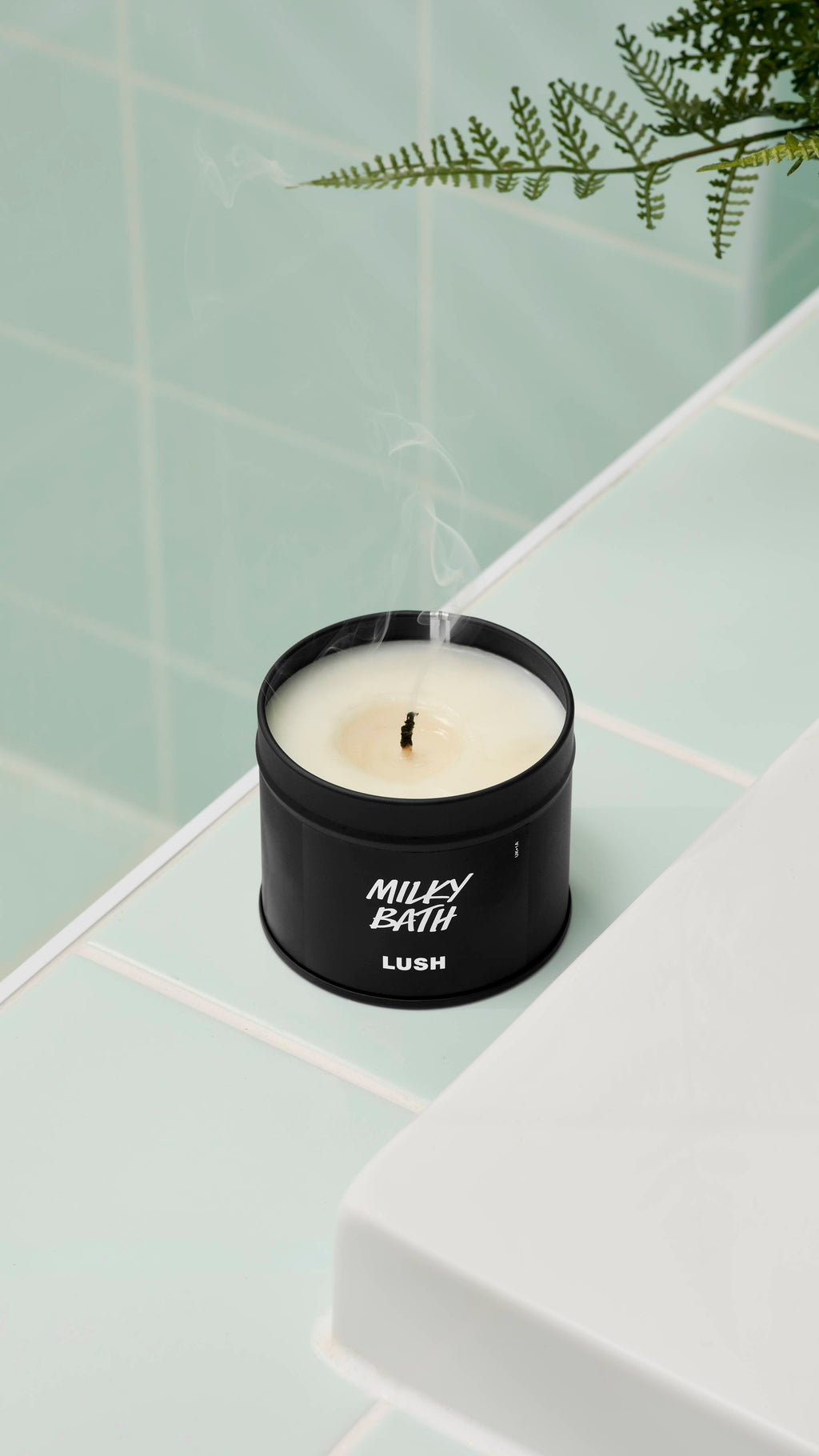 A close-up, high-angle shot of an extinguished Milky Bath candle by Lush in a matte black tin. Thin wisps of smoke rise from the blackened wick. It sits on a white ledge against mint green wall tiles with a fern leaf visible at the top.