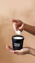 Hands hold a black pot of Lush Karma Kream body lotion against a warm beige background. One hand holds the open tub, while fingers scoop out a dollop of the rich, white cream.