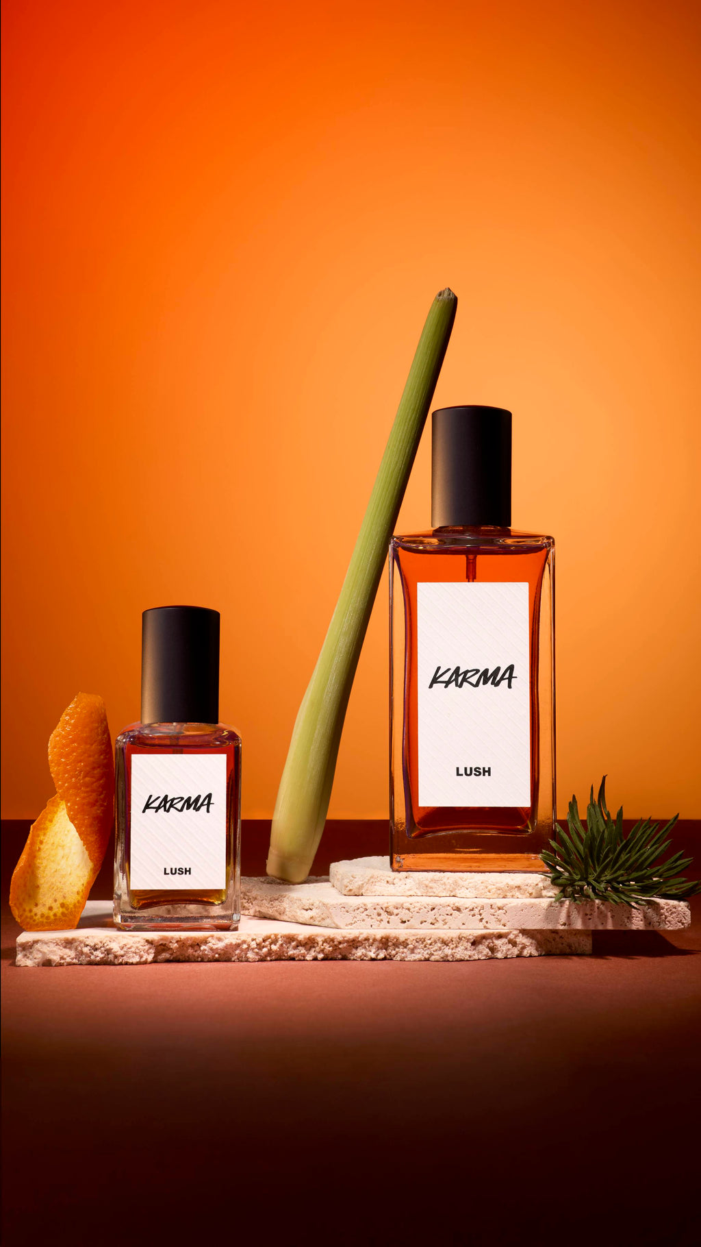 Two bottles of Lush 'Karma' perfume stand on textured stone slabs against an orange background. They are styled with a curl of orange peel, a green stalk of lemongrass, and a small sprig of pine, reflecting the scent's ingredients.