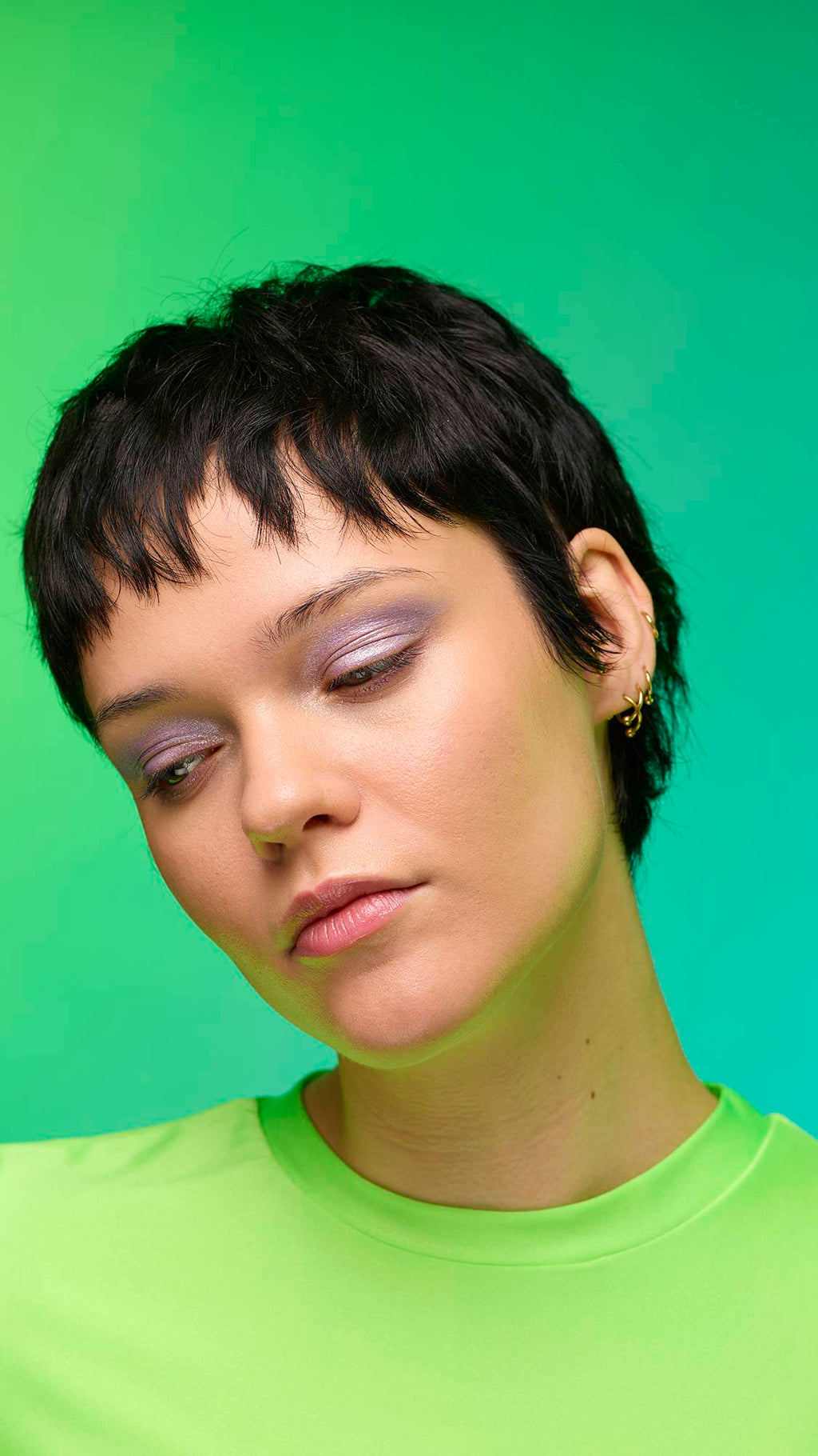 The image shows a close-up of the model's face on a pale blue-to-green gradient background. The model's make-up is done with the focal point being a delicate, purple, shimmering eyeshadow made by the Little Sparkler Glitter Crumble.