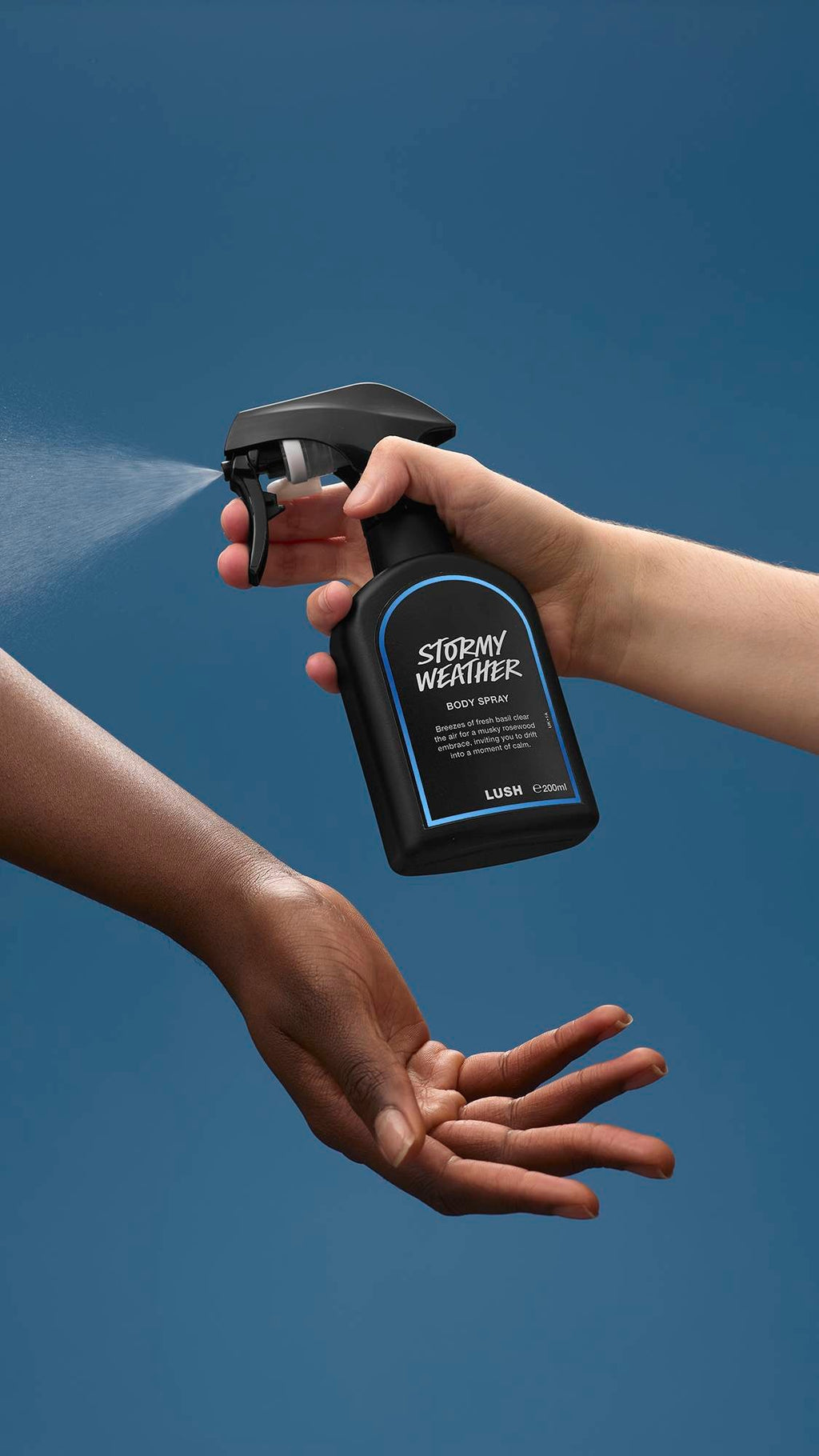 The image shows a light-toned hand spraying a bottle of LUSH 'Stormy Weather' body spray. A warm-toned hand is cupped below, catching the fine mist against a sea-blue background.