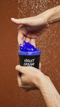 A close-up image of model scooping the vibrant Violet Cream shampoo out of the LUSH pot under running shower water.